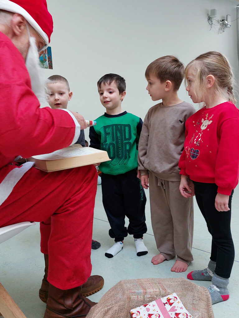Der Nikolaus hat ein grosses Buch auf den Knien liegen und liest daraus vor waehrend vier Kinder vor ihm stehen