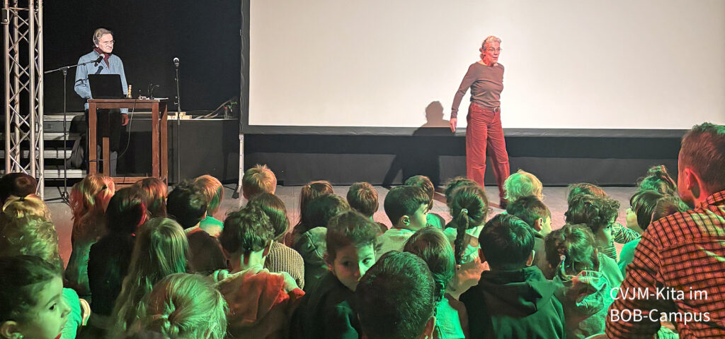 Kinder schauen zu einer Frau auf der Buehne die vor einer weissen leinwand steht