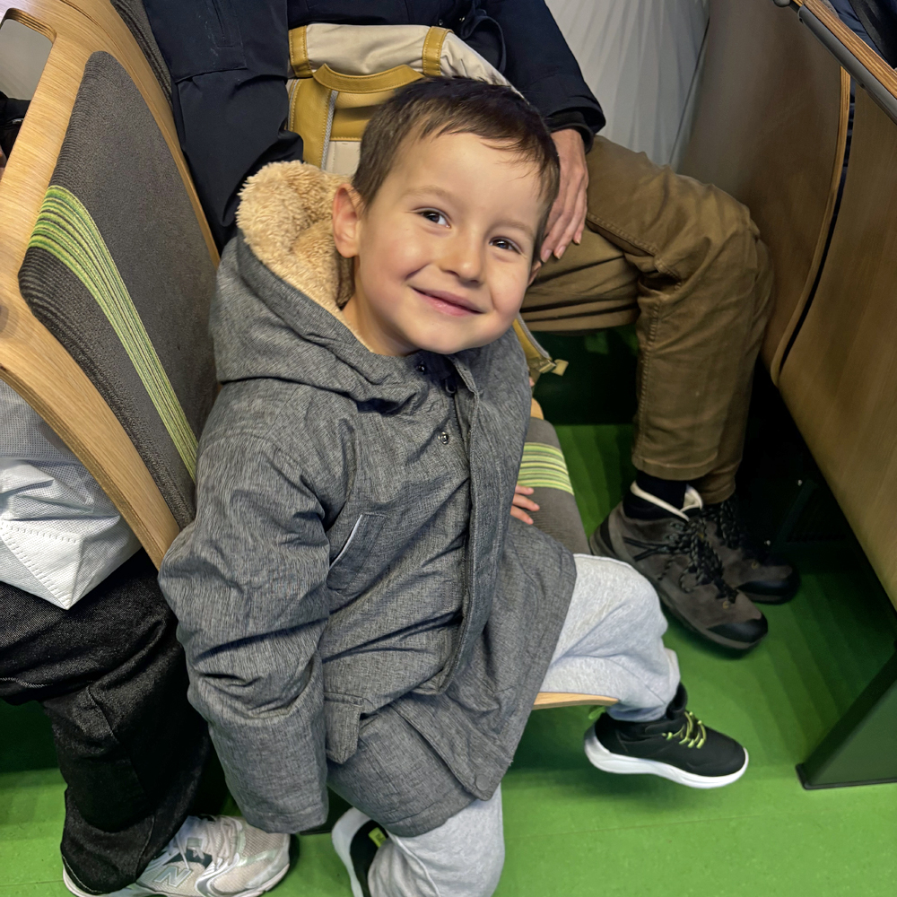 Ein Kind sitzt auf dem Stuhl in einem Schwebebahnwaggon und laechelt in die Kamera