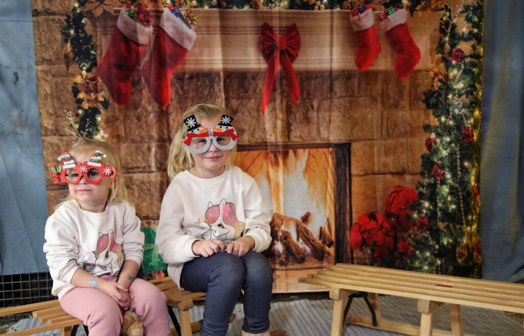 Zwei Kinder sitzen auf Schlitten vor einem Wandbehang mit Weihnachtsmotiv