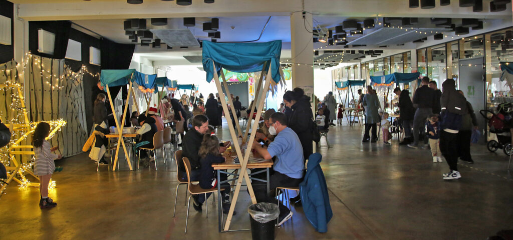 An Weihnachtsmarkt-Staenden in einer grossen Halle sitzen Menschen und sind kreativ