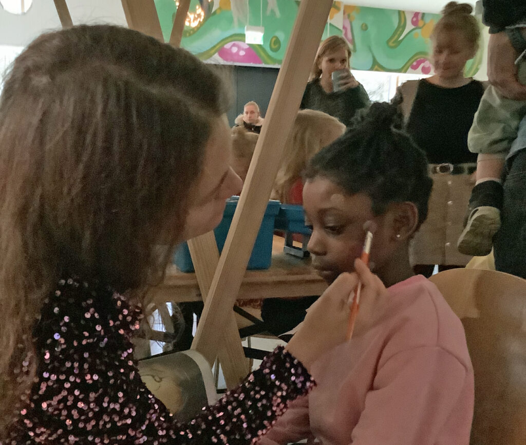 Eine Erzieherin malt an der Schminkstation mit einem Pinsel ein kleines Wintermotiv auf die Wange eines Kindes