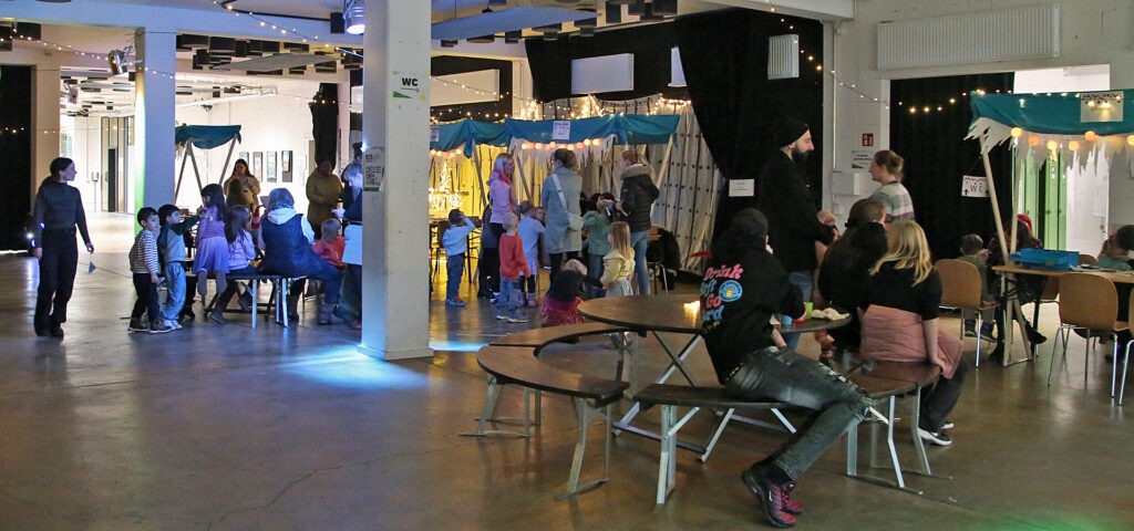 Kinder und Erwachsene stehen oder sitzen in einer Halle mit weihnachtlich geschmueckten Tischen und Baenken