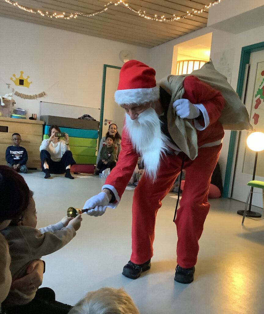 Der Nikolaus zeigt einem Kind, das im Kreis mit anderen Kindern sitzt, sein goldenes Gloeckchen