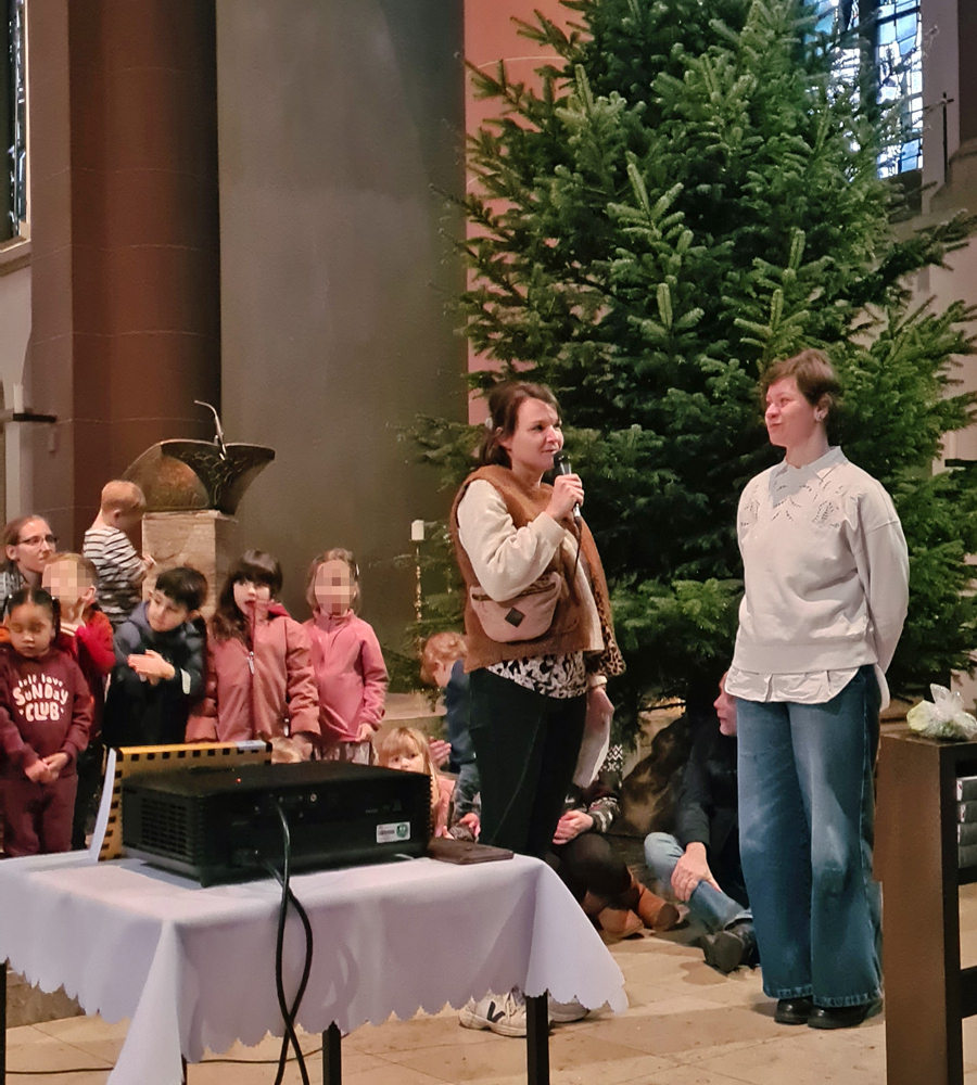 Eine Frau spricht in ein Mikrophon und schaut dabei eine andere Frau an und beide stehen in der Kirche vor einem Tannenbaum