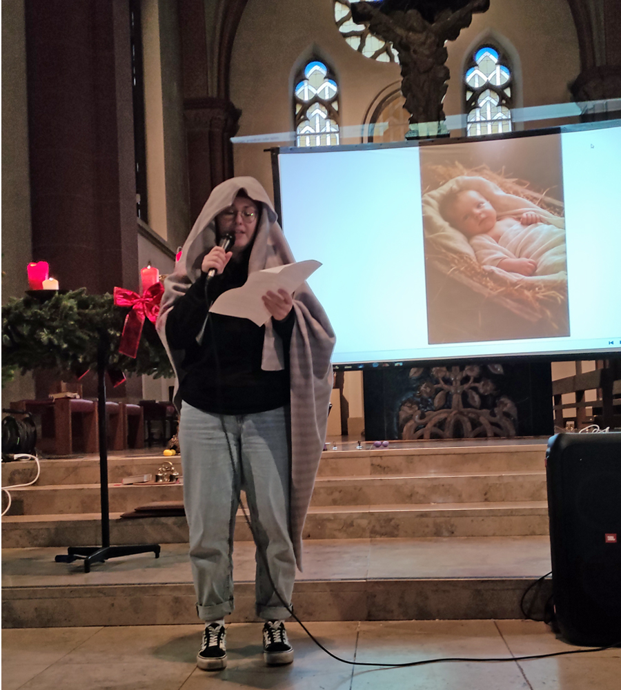 Frau hat sich eine Decke übergehängt und liest mit Mikrophon einen Text vom Blatt und hinter ihr ist auf der Leinwand ein Babyfoto zu sehen