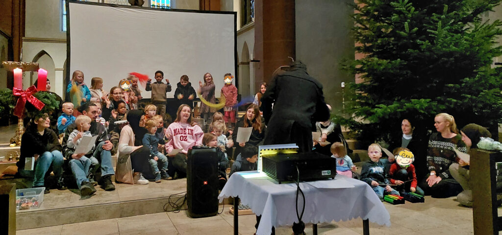 Ein Mann dirigiert vor Kindern und Erzieherinnen ein Lied mit Bewegungen in einer grossen Kirche