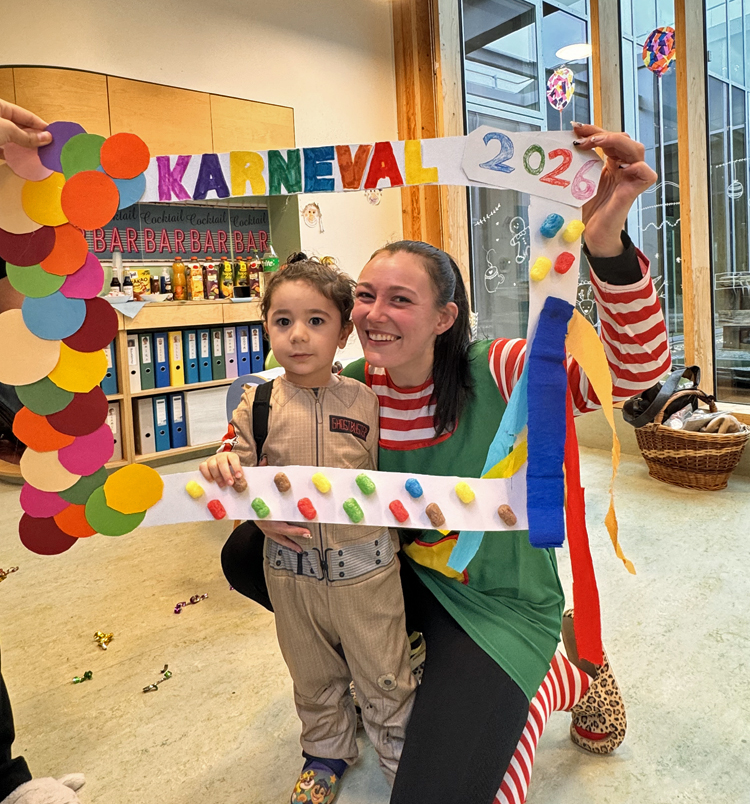 Ein Kind und eine Frau schauen durch einen bunten Papierrahmen mit der Aufschrift Karneval 2026