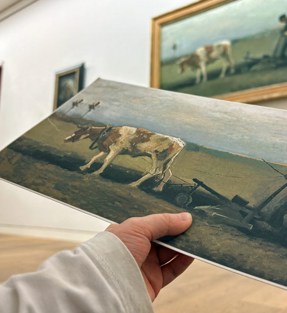 Eine Person haelt ein Bild in den Haenden mit dem gleichen Motiv eines Bildes an der Museumswand