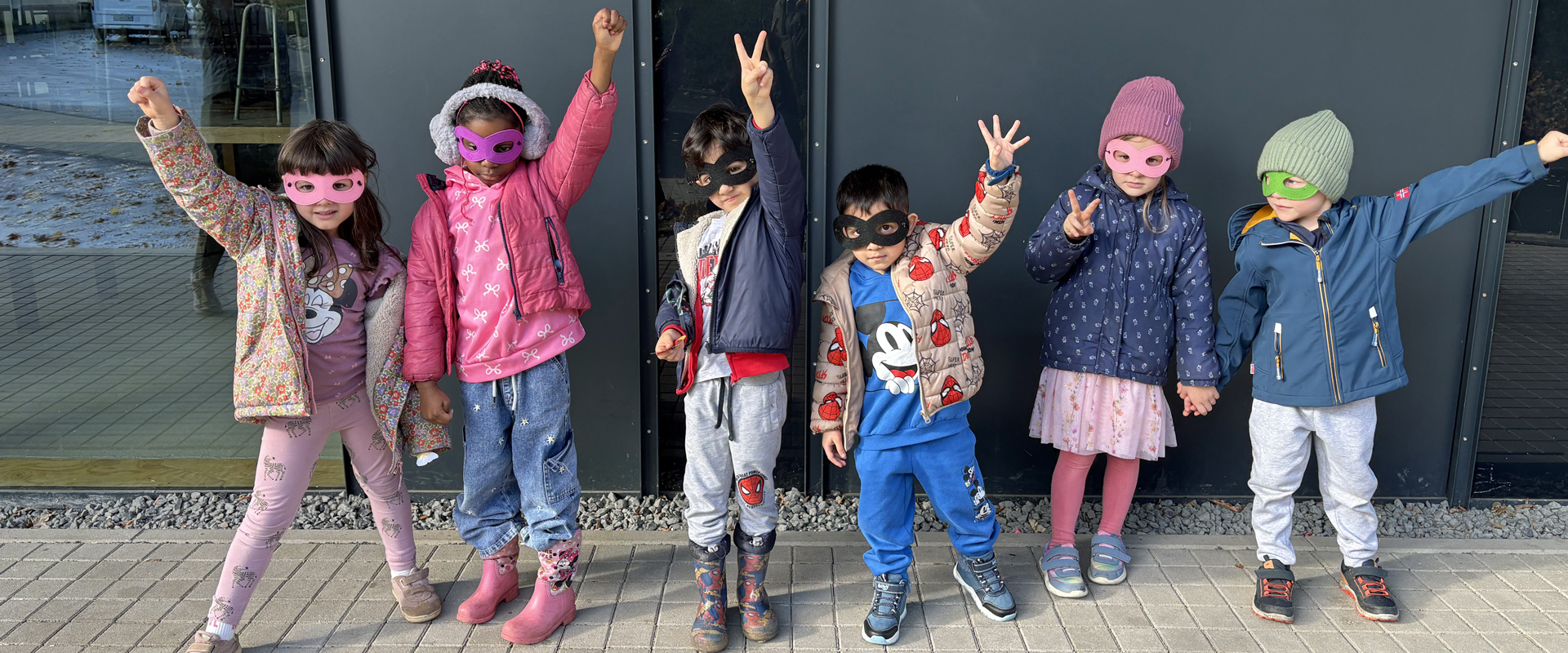 Kinder mit Augenmasken stehen vor der Kita und strecken ihre Faeuste in die Hoehe