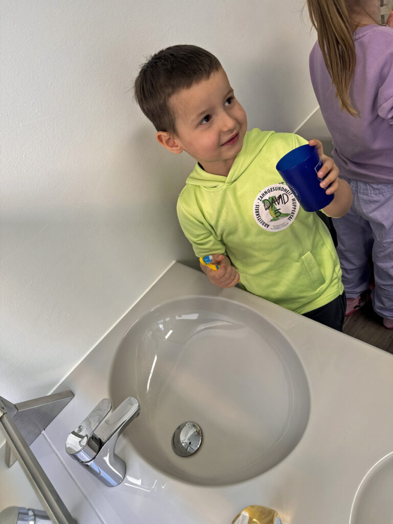 Ein Kind steht am Waschbecken und haelt Zahnbuerste und Zahnbecher in seinen Haenden