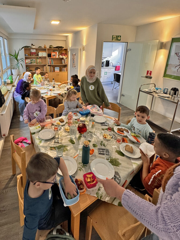 Kinder und Erwachsene sitzen an zwei Tischen und fruehstuecken