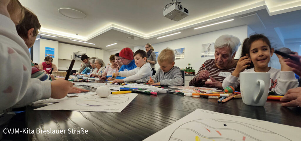 Kita-Kinder und Erwachsene sitzen am Tisch auf dem Ausmalbilder und Stifte liegen