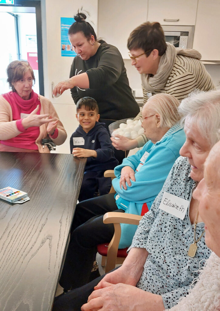 Ein Kind sitz zwsichen Seniorinnen am Tisch und es werden weise Huehnereier verteilt