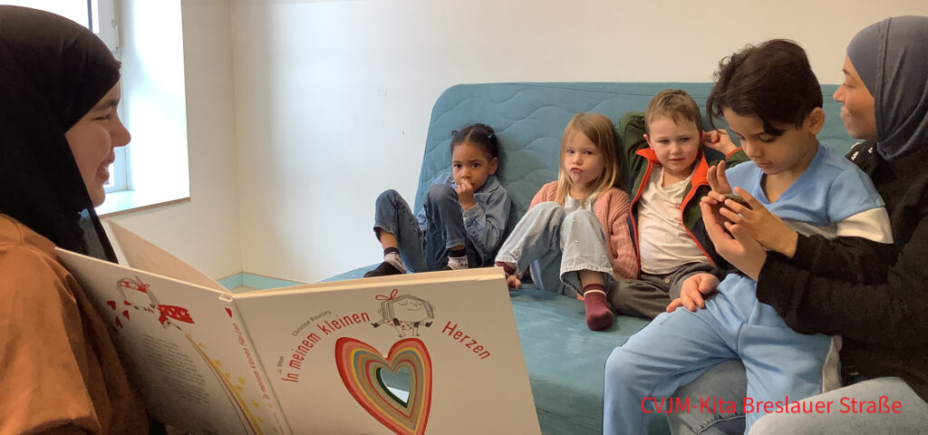 Kinder sitzen auf einem Sofa und eine Erzieherin liest ihnen aus einem Buch vor