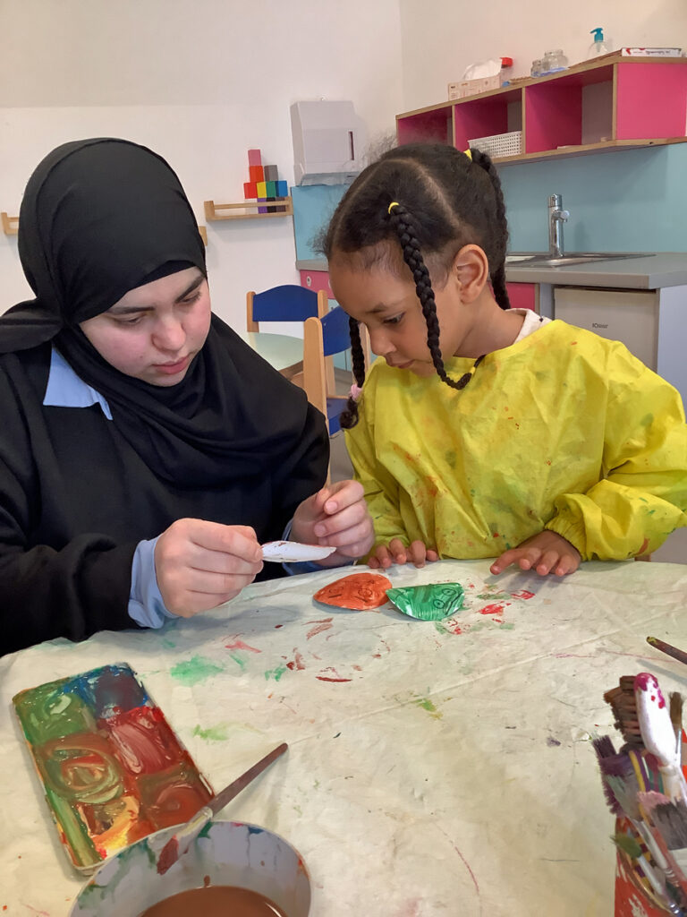 Eine Frau sitzt neben einem Kind am Tisch und sie schauen bunte Papier-Smileys an