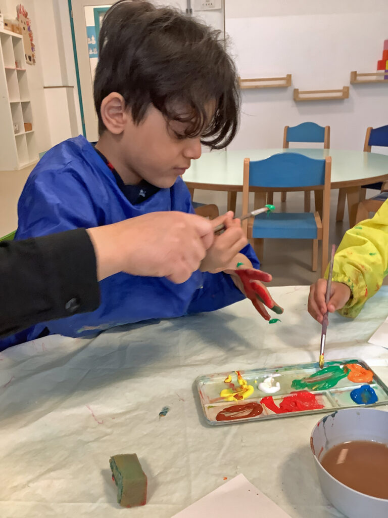 Ein Kind sitzt am Tisch und malt mit einem Pinsel seine linke Handinnenflaeche bunt an
