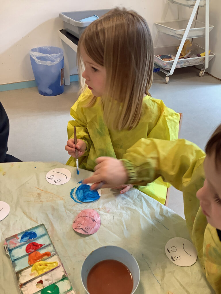 Ein Kind malt mit Wasserfarbenn ein weisses Smiley Gesicht bunt