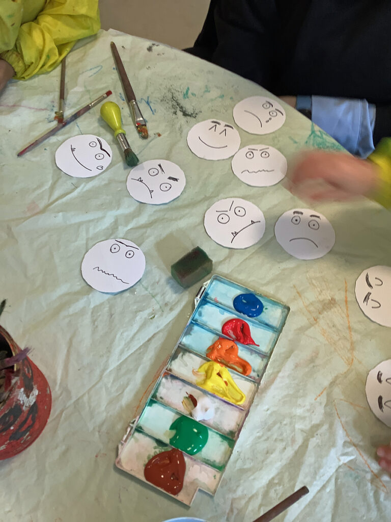 Weisse Papier-Smileys liegen auf dem Tisch neben einem Deckfarben-Kasten