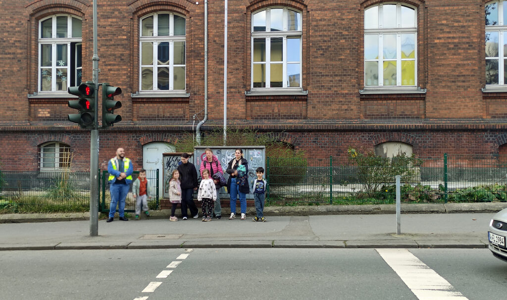 Eine Gruppe aus Kindern und Eltern steht an einer Fussgaenger-Ampel und wartet auf Gruen