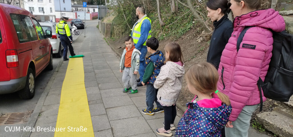 Die Verkehrspolizistin rollt ein gelbes Band auf dem Gehweg aus hinter dem die Kinder und ihre Eltern stehen