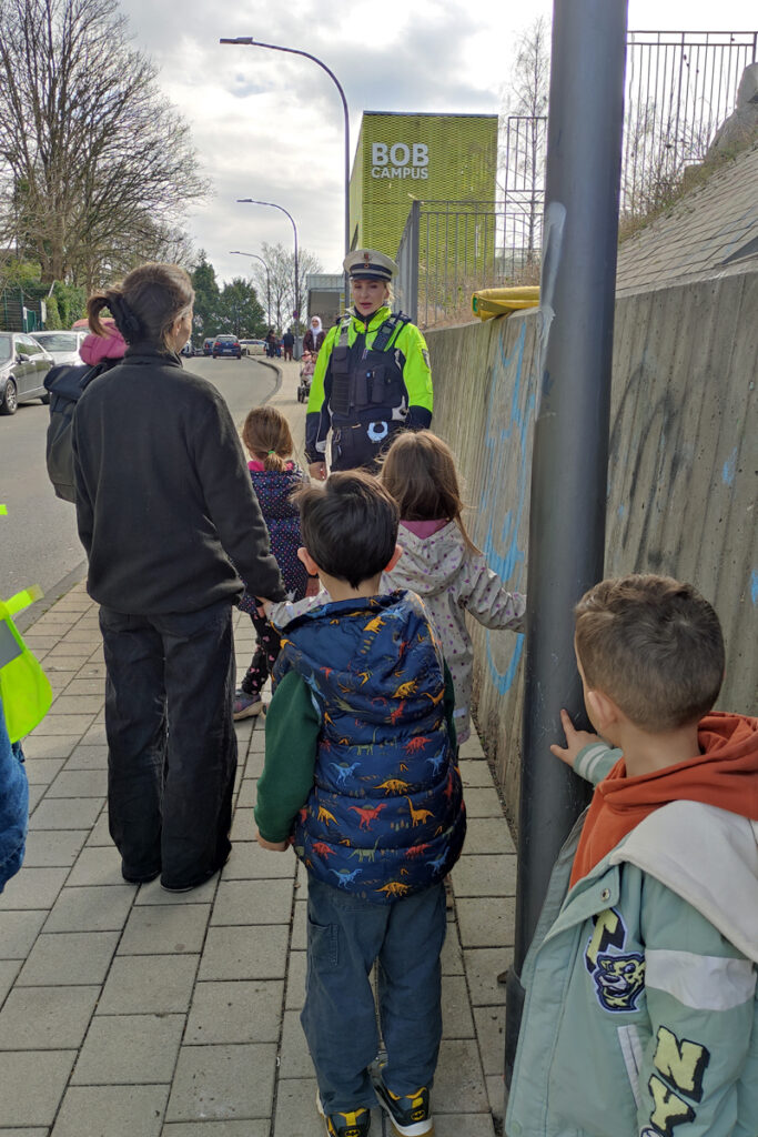 Zusammen mit einer Verkehrspolizistin geht eine Kindergruppe ueber den Gehweg zum BOB Campus
