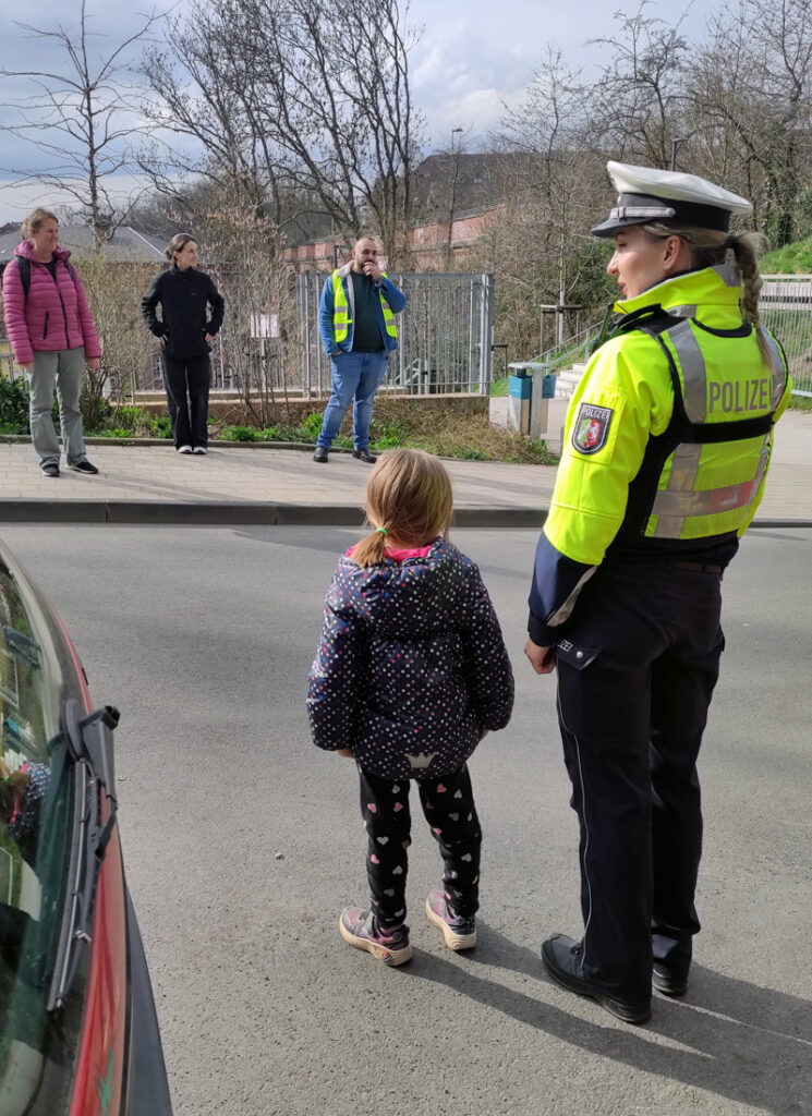 Ein Kind steht neben der Polizistin und schaut auf die Strasse und gegenueber stehen Erwachsene