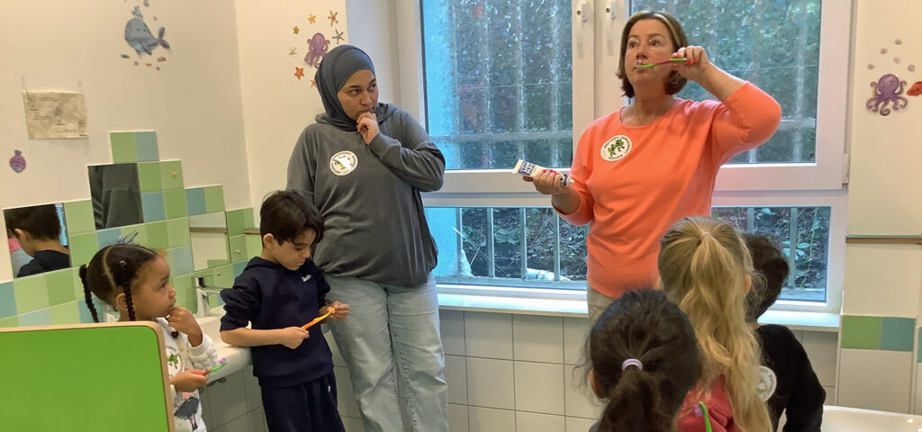 Kinder schauen einer Erwachsenen zu die ihnen das richtige Zaehneputzen zeigt