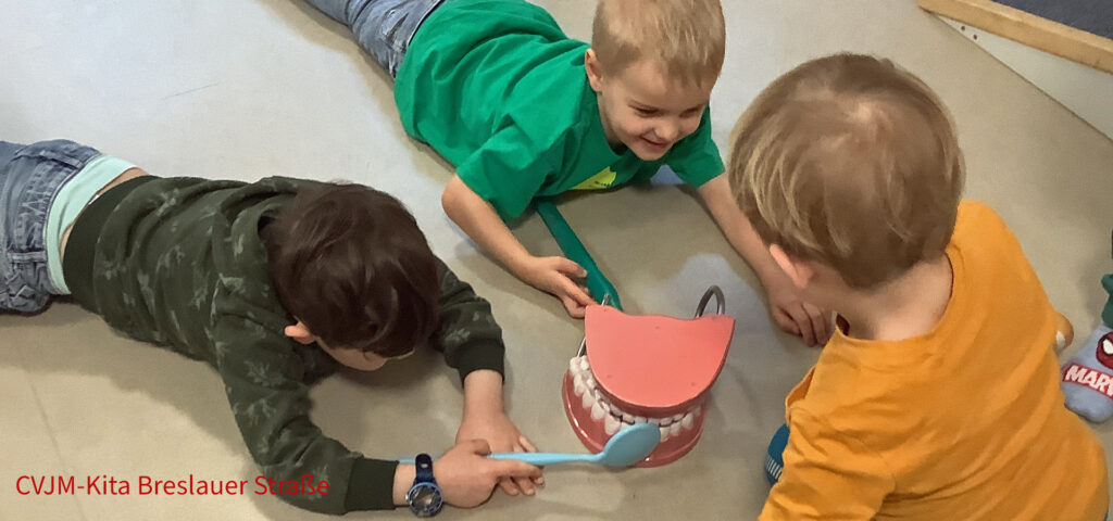 Kinder liegen auf dem Bauch um ein grosses kuenstliches Gebiss herum und untersuchen dessen Zaehne