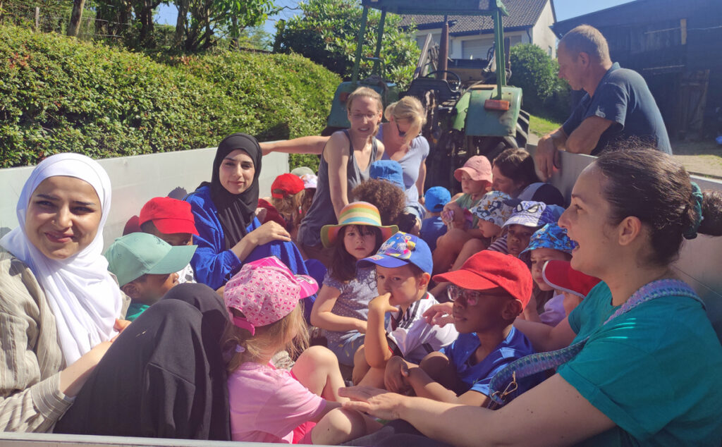 Kinder und Erzieherinnen sitzen bei Sonnenschein im Anhaenger eines Traktors