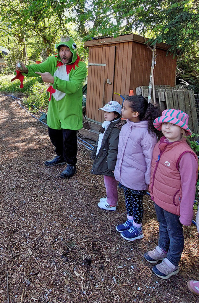 Kinder schauen zu einem erwachsenen Mann in Froschkostuem der ihnen einen Frosch zeigt