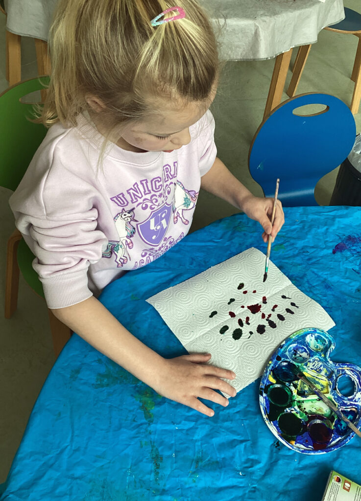 Ein Kind sitzt am Tisch und tupft Farbe mit einem Pinsel auf ein Papiertuch