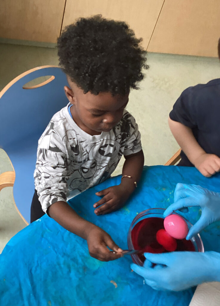 Ein Kind sitzt am Tisch und taucht weisse Eier mit einem Loeffel in ein rotes Farbbad