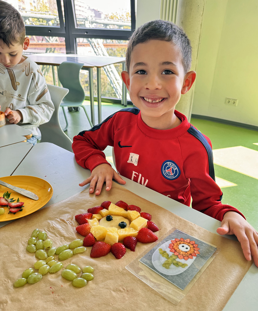 Ein Kind sitzt am Tisch und vor ihm liegt auf Backpapier ein Blume aus Weintrauben, Erdbeeren und Ananasstuecken