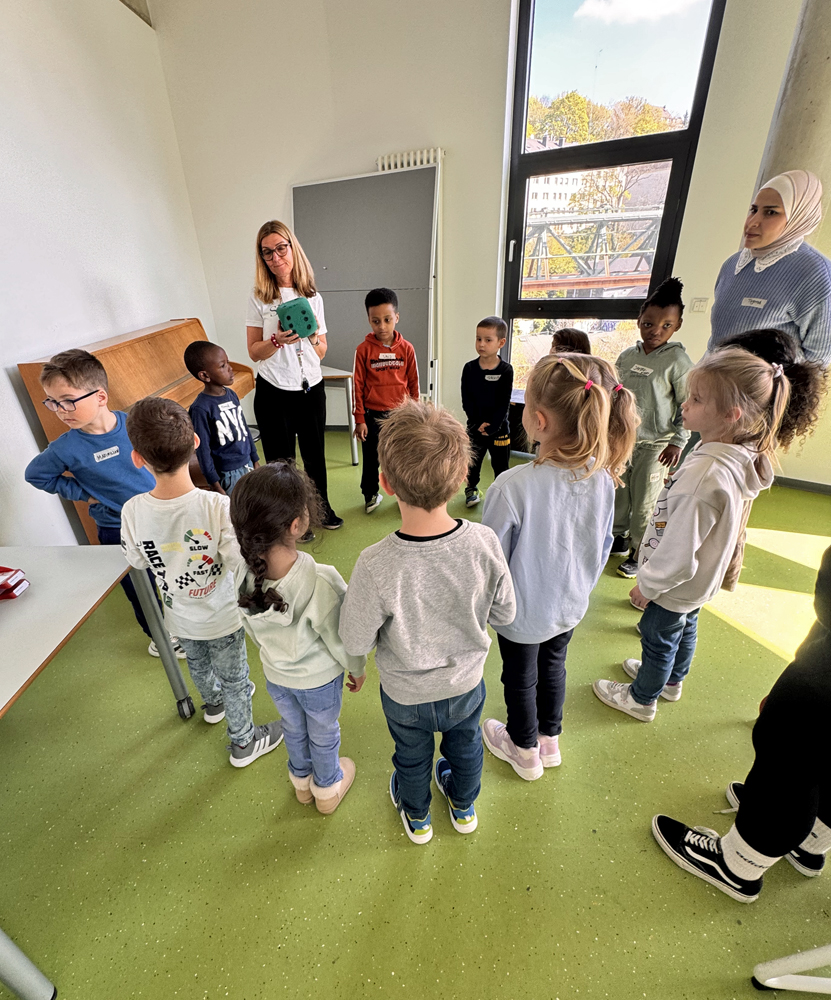 Kinder und zwei Frauen stehen im Kreis in einem Raum der Junioruni