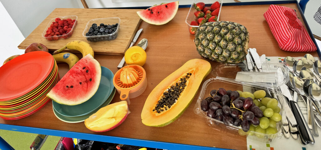 Auf einem Tisch liegen viele verschiedene Fruechte, bunte Teller, Messer und kleine Loeffel