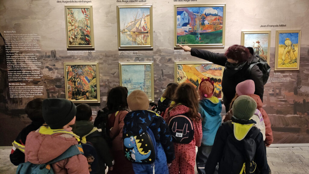 Eine Frau zeigt auf Bilder von Vincent van Gogh und erklaert einer Kindergruppe etwas