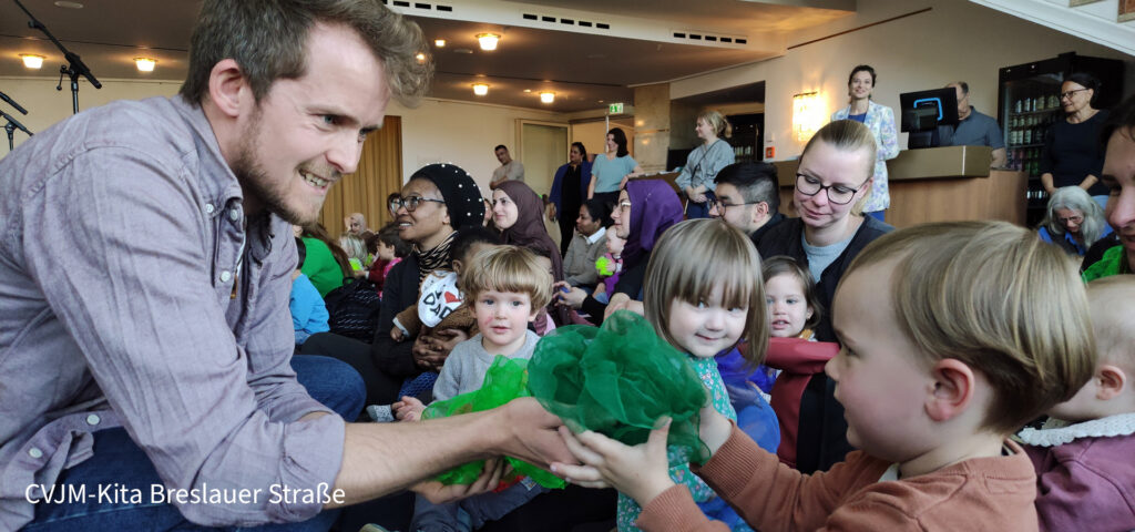 Ein Mann ueberreicht einem Kind im Opernfoyer Wuppertal gruene Tuecher und andere Kinder schauen zu