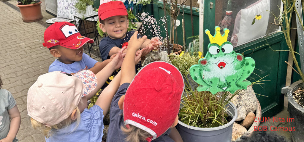 Kinder halten ihre Haende über einen kleinen Springbrunnen, der aus einem grossen Blumenkuebel sprudelt