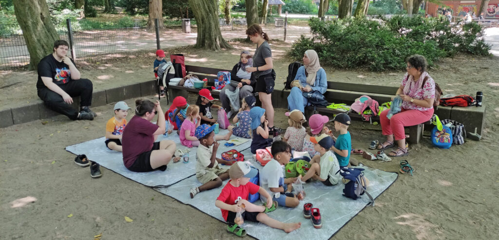Kinder sitzen auf einer Picknickdecke auf einem Spielplatz und essen zu Mittag