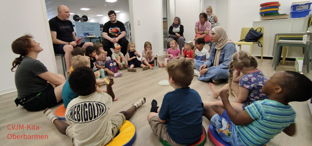 Kinder sitzen im Kreis auf Kissen und hoeren einer Erzieherin zu