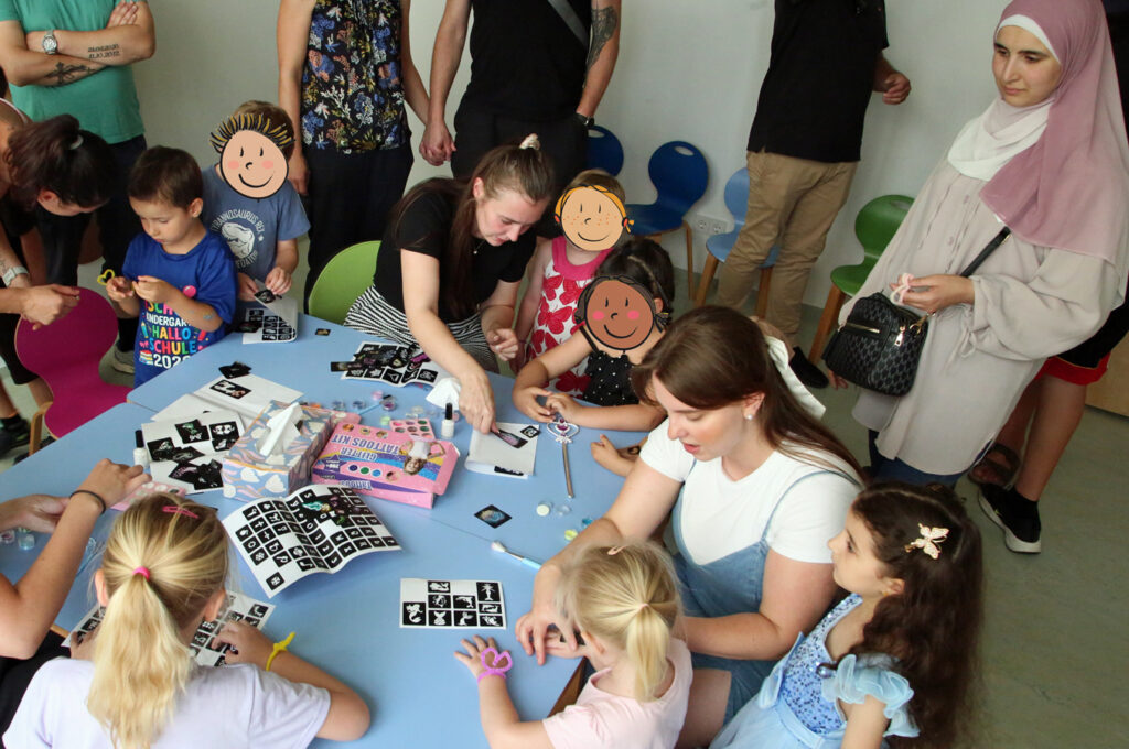 Kinder und Erzieherinnen sitzen am Tisch auf dem das Equipment fuer Glitzertattoos liegt