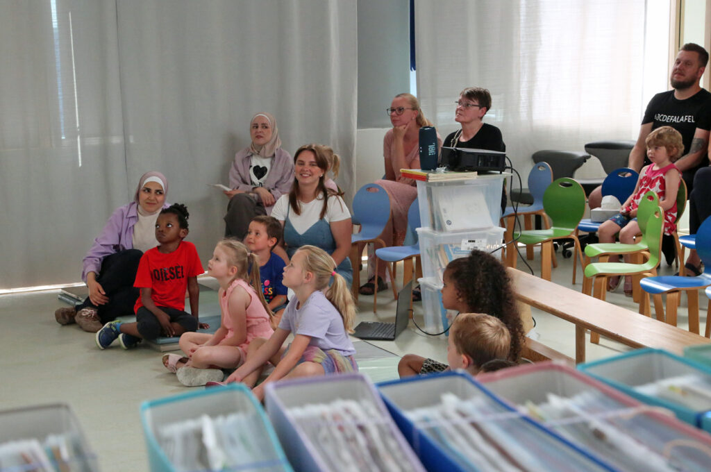 Kindern hocken auf dem Boden, Erwachsene sitzen auf kleinen Stuehlen und alle schauen einen Film
