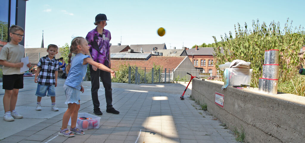 Ein Kind wirft einen Ball auf gestapelte Dosen und weitere Kinder stehen hinter ihm und warten