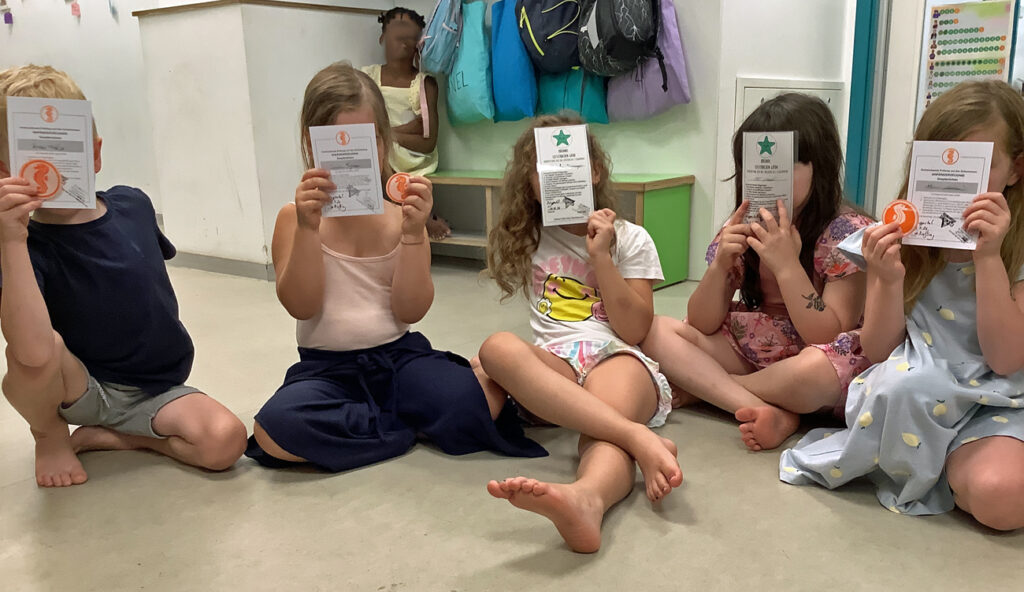 Kinder hocken auf dem Fussboden und zeigen ihre Seesternchen und Seepferdchen Urkunden