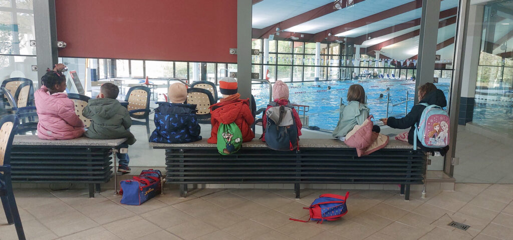 Kinder sitzen im Flur des Hallenbades Langerfeld und schauen durch eine Scheibe in das Schwimmerbecken