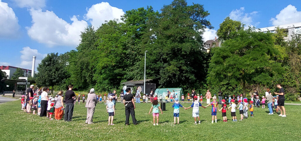 Die groesseren Kita-Kinder stehen mit Erwachsenen auf einer Wiese im Kreis und halten sich an den Haenden