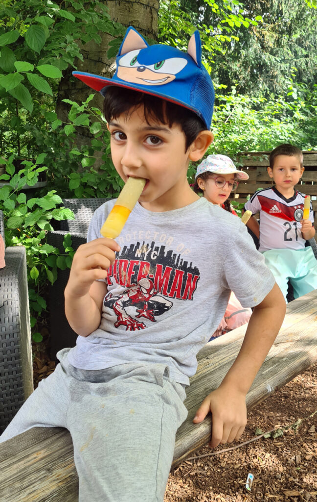 Kinder sitzen draussen in der Natur auf Holzbaenken und essen Eis