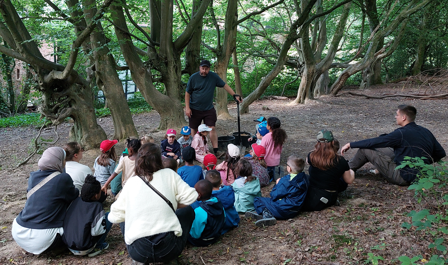 Kinder und Erwachsene sitzen im Wald im Kreis und hoeren dem Waldpaedagogen zu