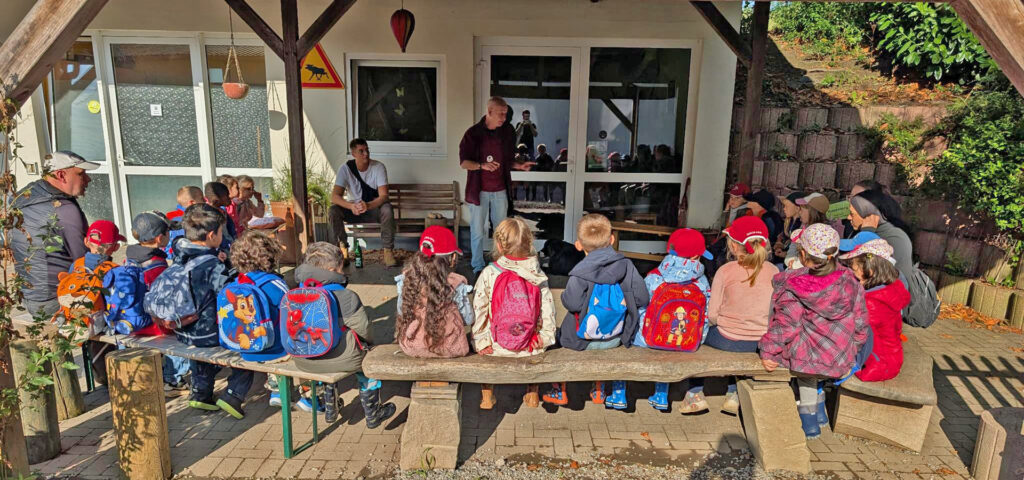 Die Vorschulkinder sitzen auf Holzbaenken vor einem Haus und hoeren einem Mann zu